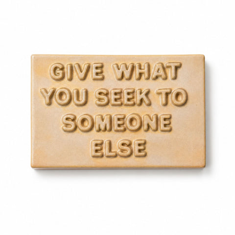A rectangular ceramic sign with the text reading 'GIVE WHAT YOU SEEK TO SOMEONE ELSE' in a warm beige and light brown glaze, shown on a white background.