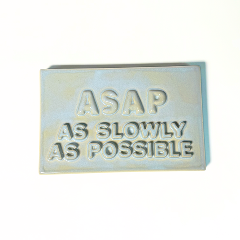 A rectangular ceramic sign with text reading 'ASAP AS SLOWLY AS POSSIBLE' in a light blue-gray glaze on a white background.
