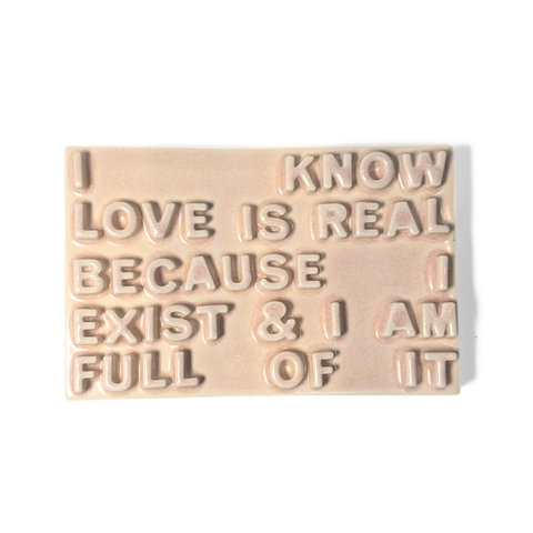 A rectangular peach ceramic sign with raised lettering that reads 'I KNOW LOVE IS REAL BECAUSE I EXIST & I AM FULL OF IT' on a plain white background.