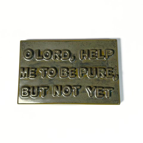 A rectangular dark green-colored ceramic sign with the text reading 'O LORD, HELP ME TO BE PURE, BUT NOT YET' on a plain white background.