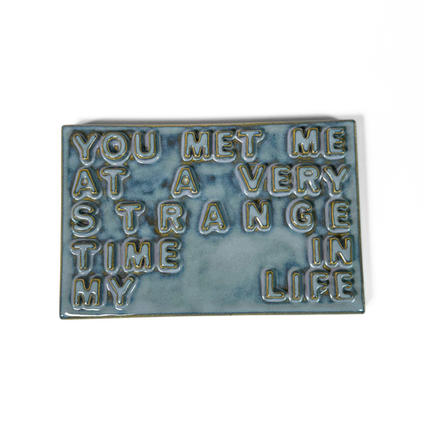 A rectangular ceramic sign with raised, textured lettering that reads 'YOU MET ME AT A VERY STRANGE TIME IN LIFE' in a light blue-gray glaze with gold-toned outlines, shown on a plain white background.