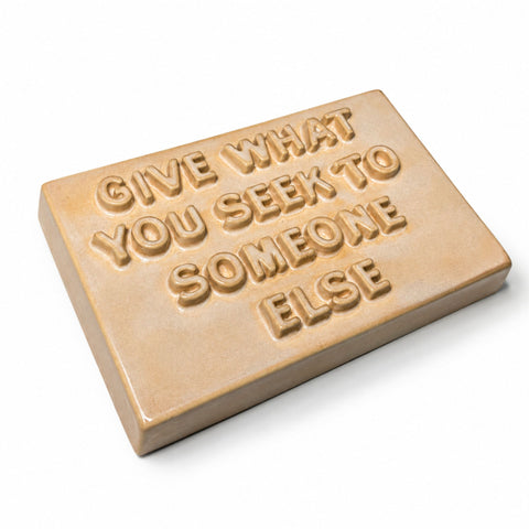 A rectangular ceramic sign with the text reading 'GIVE WHAT YOU SEEK TO SOMEONE ELSE' in a warm beige and light brown glaze, shown on a white background.