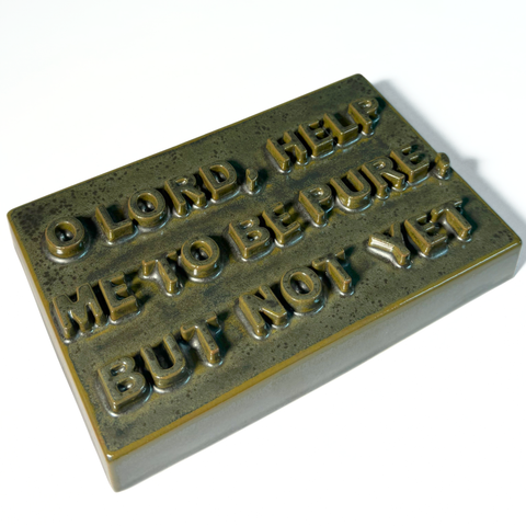 A rectangular dark green-colored ceramic sign with the text reading 'O LORD, HELP ME TO BE PURE, BUT NOT YET' on a plain white background.