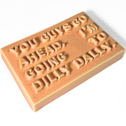 A rectangular ceramic sign with a warm orange-peach glaze, featuring raised text that reads 'YOU GUYS GO AHEAD. I'M GOING TO DILLY DAILY.