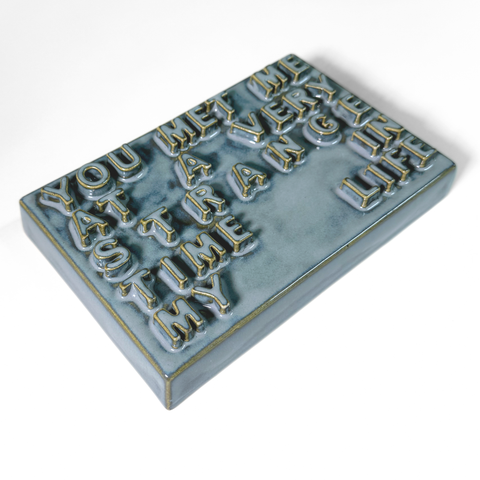 A rectangular ceramic sign with raised, textured lettering that reads 'YOU MET ME AT A VERY STRANGE TIME IN LIFE' in a light blue-gray glaze with gold-toned outlines, shown on a plain white background.