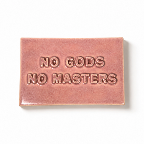 A rectangular pink ceramic sign with the text 'NO GODS NO MASTERS' in all caps, shown on a plain white background.