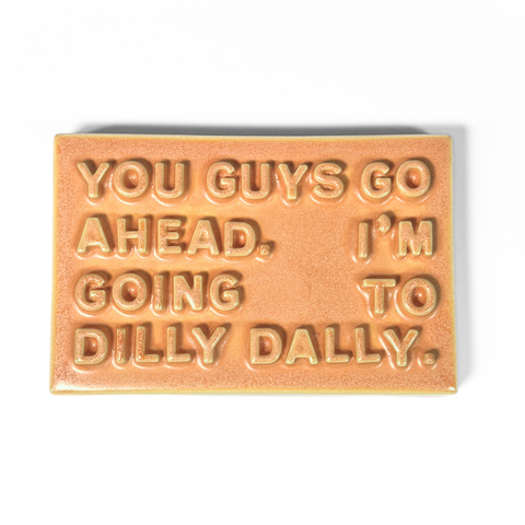A rectangular ceramic sign with a warm orange-peach glaze, featuring raised text that reads 'YOU GUYS GO AHEAD. I'M GOING TO DILLY DAILY.