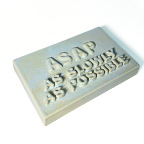A rectangular ceramic sign with text reading 'ASAP AS SLOWLY AS POSSIBLE' in a light blue-gray glaze on a white background.