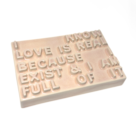 A rectangular peach ceramic sign with raised lettering that reads 'I KNOW LOVE IS REAL BECAUSE I EXIST & I AM FULL OF IT' on a plain white background.